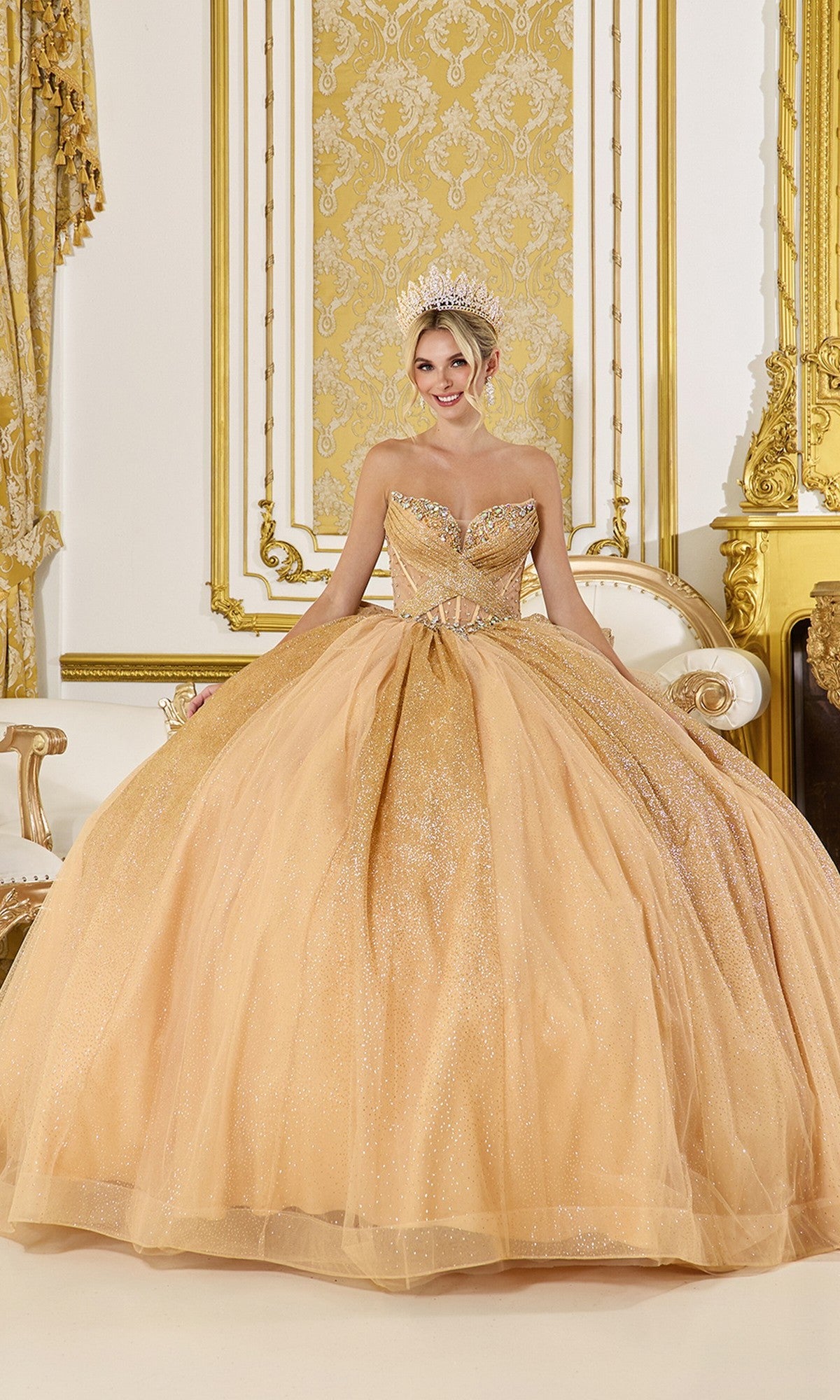 A woman in the Quinceanera Dress 5078 By Dancing Queen in gold, with a lace-up back and tiara, sits on an ornate white and gold chair in a lavishly decorated room with gold accents.